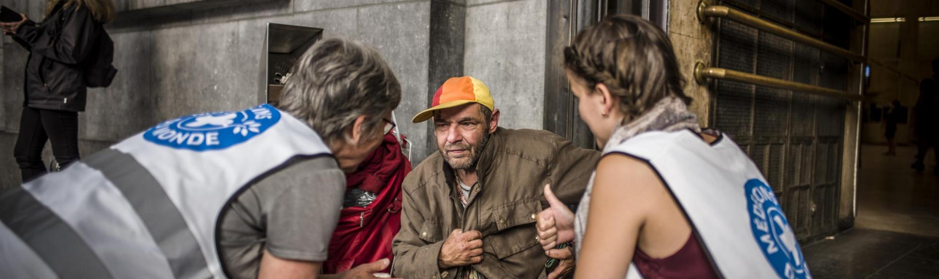Devant la gare centrale de Bruxelles, deux travailleuses de Médecins du Monde sont assises aux côtés d'un homme sans domicile fixe, lui expliquant leur mission.