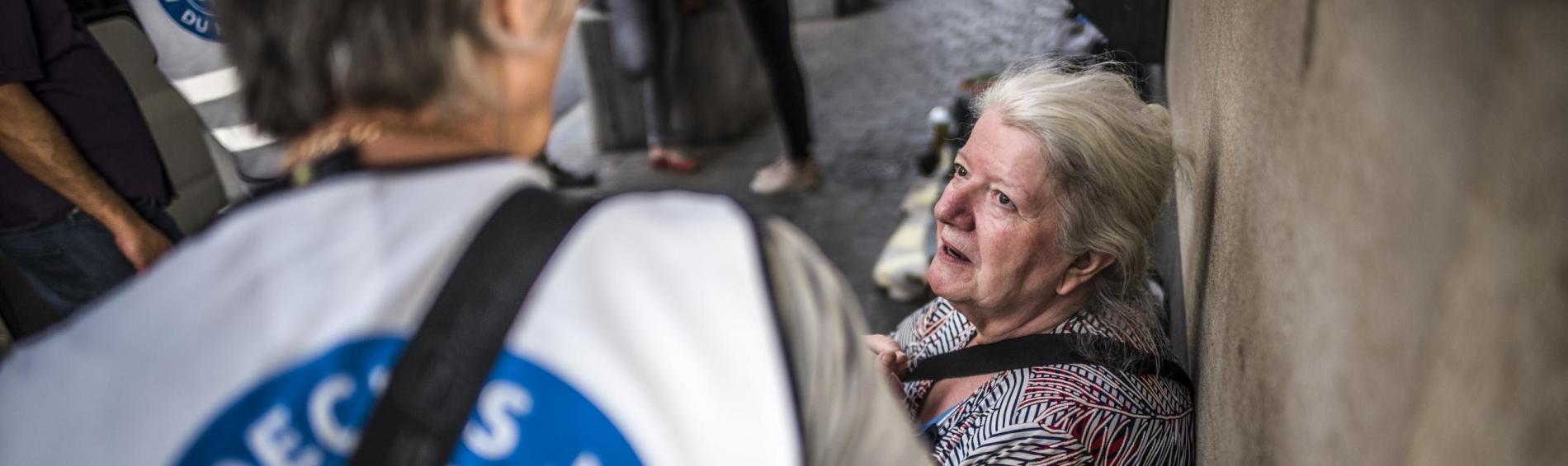 Devant la Gare Centrale de Bruxelles, juste devant le Médibus de Médecins du Monde. Une dame aux longs cheveux blancs, dressés en chignon, est assise sur un banc avec une bénévole de Médecins du Monde, vue de dos.