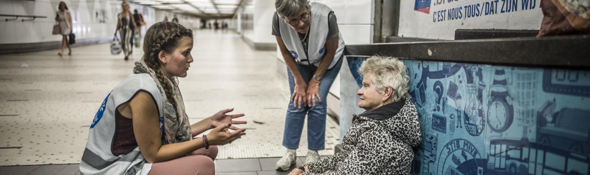 Dans le couloir d'une station de métro, deux travailleuses de Médecins du Monde discute avec une dame agée, assise sur le sol, sur une feuille de journal. Cheveux courts et blancs, elle porte un épais manteau à pois sur une robe rose arrivant à ses genoux, et des chaussures de marche.