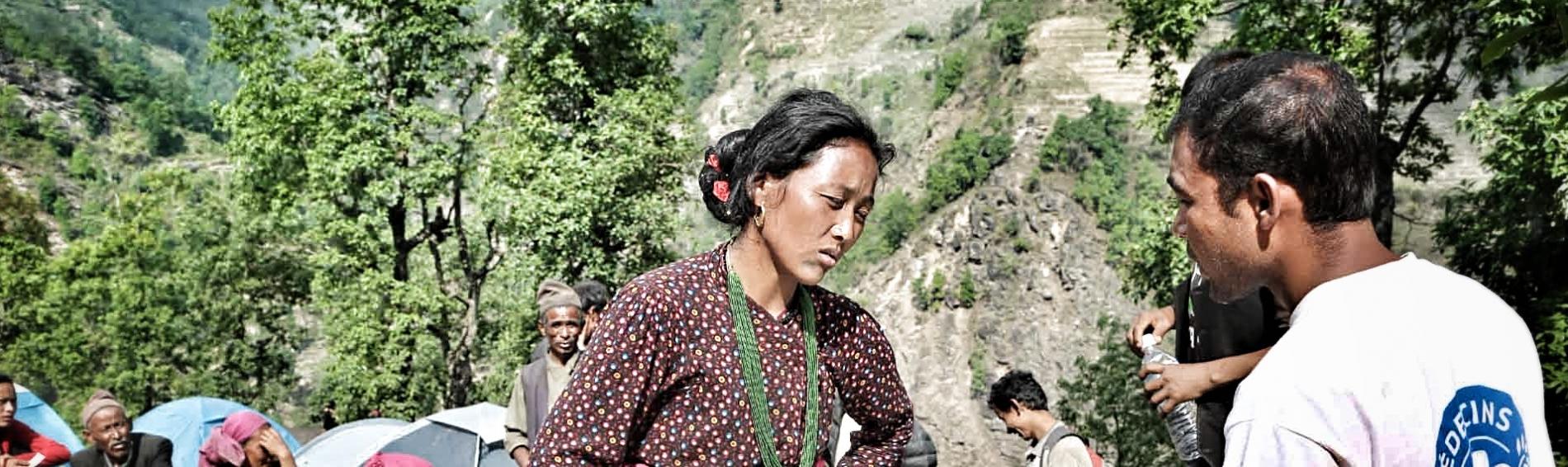 Dans un décor montagneux, un travailleur de Médecins du Monde discute avec une femme dont les mains sont posées sur son ventre. En arrière plan, des tentes et des personnes assises autour.