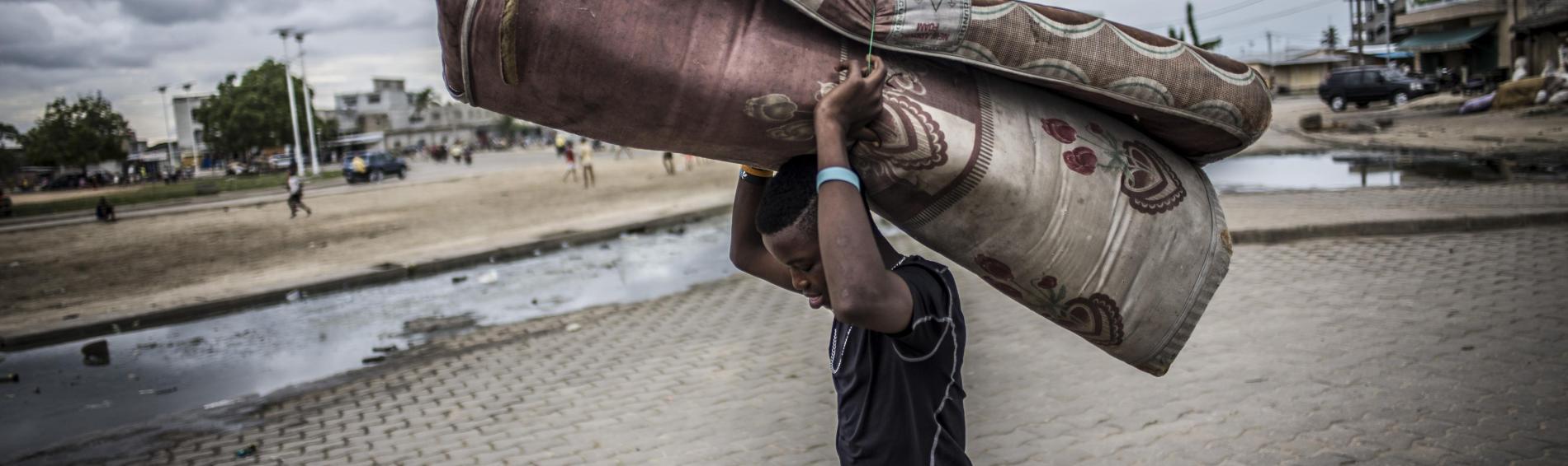 Un adolescent dans la rue au Bénin. Le temps est nuageux, il porte un matelas humide et bien plié sur la tête. À l'arrière-plan, des enfants jouent dans une banlieue déserte.