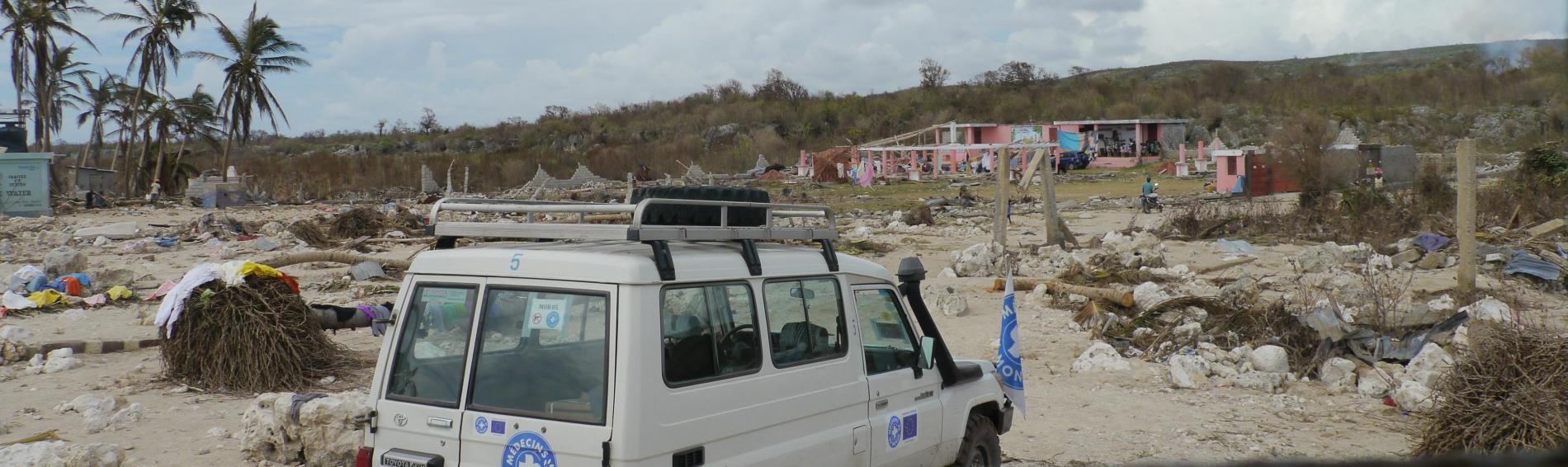 À Haïti, sur une place, une voiture de Médecins du Monde est garée devant un paysage de catastrophe : les maisons sont presque à terre, les toits envolés, les débris jonchent le sol.
