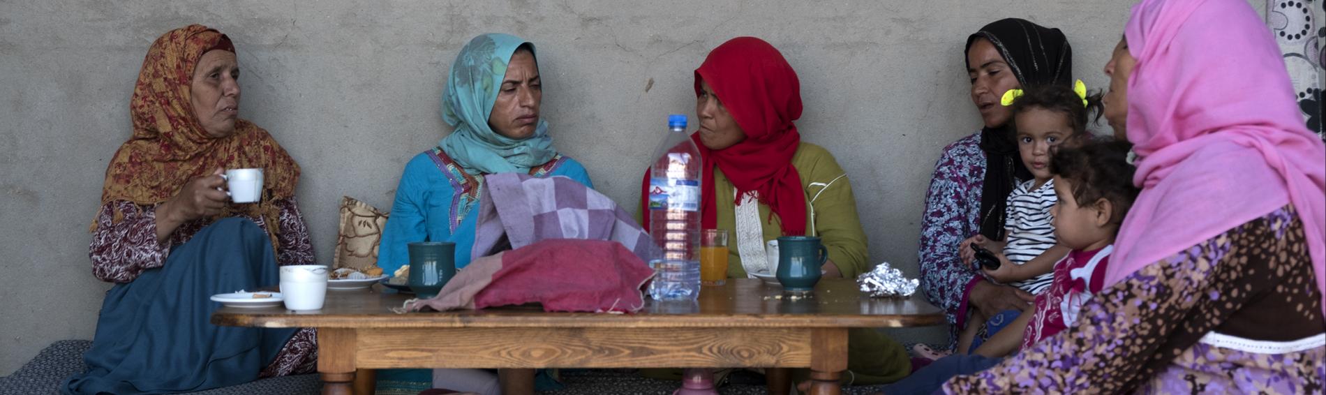 Cinq femmes à l'extérieur d'une maison, en Tunisie, en habits colorés. Elles sont assises autour d'une table sur laquelle se trouve se l'eau et du café.