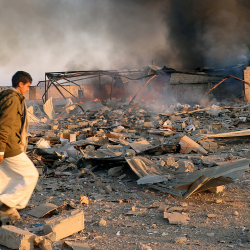 Dans une paysage de ruines, de fumée et de flammes, un jeune homme marche dans les décombres.