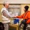 Dans un cabinet médical, un médecin portant la chasuble Médecins du Monde, souriant, sert la main d'une jeune homme portant une grosse veste orange et un bonnet, après une consultation.